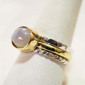 May include: A silver and gold ring with a large, round, white moonstone. The ring is made of three bands, one with a braided design, one with a hammered texture, and one with a smooth finish.