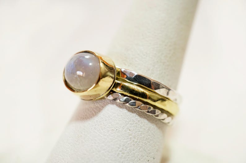 May include: A silver and gold ring with a large, round, white moonstone. The ring is made of three bands, one with a braided design, one with a hammered texture, and one with a smooth finish.