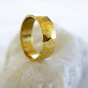May include: A gold hammered ring with a wide band. The ring is sitting on a white rock.