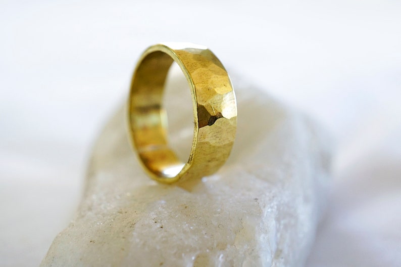 May include: A gold hammered ring with a wide band. The ring is sitting on a white rock.