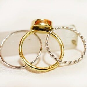 May include: Three gold rings stacked on top of each other. The top ring has a large, round, gold stone. The bottom two rings are silver and have a twisted design.