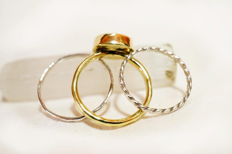 May include: Three gold rings stacked on top of each other. The top ring has a large, round, gold stone. The bottom two rings are silver and have a twisted design.