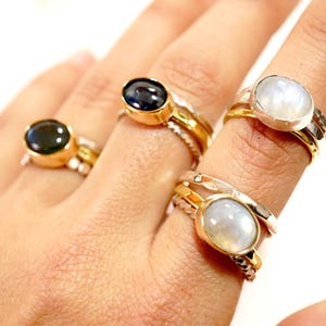 May include: Three gold rings with oval-shaped gemstones. The rings are stacked on a finger. The gemstones are a dark blue, a white moonstone, and another white moonstone.