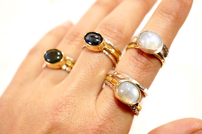 May include: Three gold rings with oval-shaped gemstones. The rings are stacked on a finger. The gemstones are a dark blue, a white moonstone, and another white moonstone.