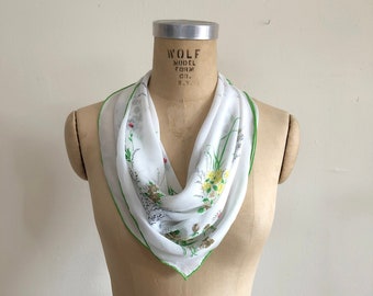 May include: A white square scarf with a floral pattern and a green border is draped on a mannequin. The scarf features yellow, red, and green floral designs. The mannequin is a light tan color.