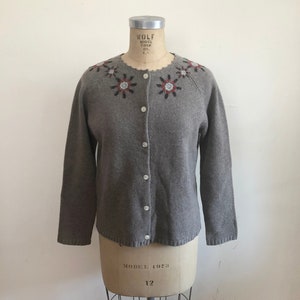 Op de afbeelding: Een grijze cardigan met een ronde hals en knoopsluiting. De trui heeft een geborduurde bloemenversiering rond de hals in tinten rood, wit en blauw.
