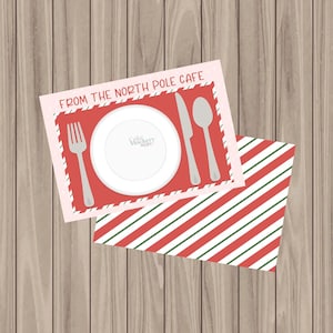 May include: A festive placemat with a red border and the text "FROM THE NORTH POLE CAFE" surrounds a white plate, fork, knife, and spoon. A second placemat with red, white, and green stripes is below. The background is a light brown wood grain.