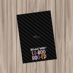 以下が含まれることがあります： 黒いカードに斜めのストライプと、紫、オレンジ、白で「You've been La-BOO BOO-ED」という文字が書かれています。カードは木製の表面にあります。「Cake Mockery」という言葉が左上にあります。