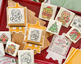 May include: A set of Christmas cookies decorated with paint-your-own designs. The cookies are shaped like bears, Christmas trees, and presents. The cookies are on a red tray with a wooden cutting board and a note that says "I'm here from the North Pole with an extra special treat! A cookie you can paint and then get to eat!"