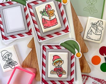May include: Christmas cookie decorating kit with elf and gift bag designs. The cookies are on a white plate with red and white striped borders. Includes stencils, a pink cookie cutter, and a card that says "A COOKIE you can PAINT, and then get to EAT!"