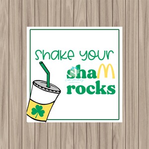 May include: A white square sign with green and yellow accents. The sign says "Shake your shamrocks". A green and yellow cartoon milkshake cup with a green straw is in the upper left corner.