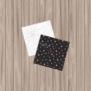 May include: Two square paper designs on a wooden surface. One features a white spiderweb pattern, and the other has a black background with a pattern of pink, gold, and white stars. The text "Cake Mocker" is visible on the black design.