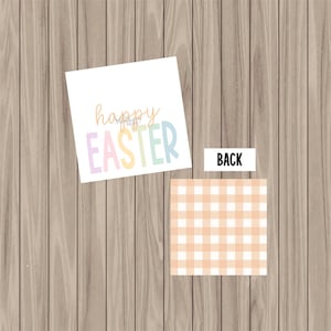 May include: A white card with the words "happy Easter" in pastel colors. The back of the card features a peach and white gingham pattern. The card is on a wooden surface.
