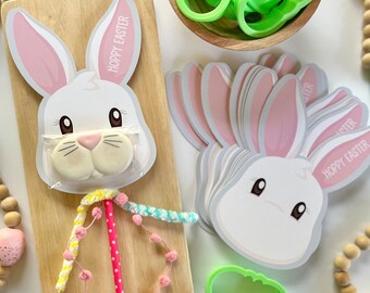 May include: A set of white bunny face party favours with pink ears and the text "Hoppy Easter". The bunny faces are stacked on top of each other. There is a wooden chopping board with a white bunny face lollipop with a pink, yellow, and blue striped stick. There are green cookie cutters in a wooden bowl.