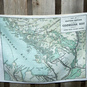 May include: A white linen tea towel displaying a vintage map of the Eastern Section of Georgian Bay, with text indicating the Grand Trunk Railway System. The map is in shades of blue and green, held by wooden pegs.