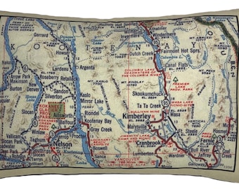 May include: Rectangular pillow featuring a vintage map design of the Kootenay region in British Columbia, Canada. The map includes roads, towns, and geographical features in shades of beige, blue, and red. The pillow has a beige border.