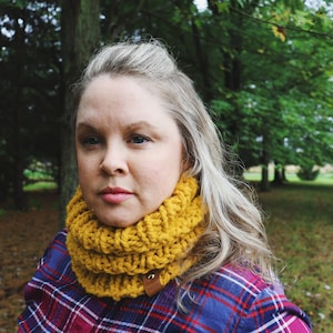 May include: A woman wearing a yellow knitted infinity scarf with a brown leather button. The scarf is thick and chunky, and the woman is wearing a plaid shirt.