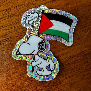 Op de afbeelding: Een glitterende sticker met een cartoonhond die een Palestijnse vlag vasthoudt. De vlag is rood, wit, zwart en groen. Een tekstballon boven de hond zegt "Free Palestine."