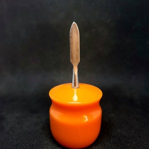以下が含まれることがあります： 銀色の金属製の蓋が付いた小さなオレンジ色の陶器の壺。蓋は尖った槍の形をしています。