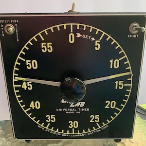May include: A black Universal Lab timer with a yellow dial and numbers. The timer has a knob in the center and an on/off switch. The text "Universal Timer Model 160" is visible on the face.