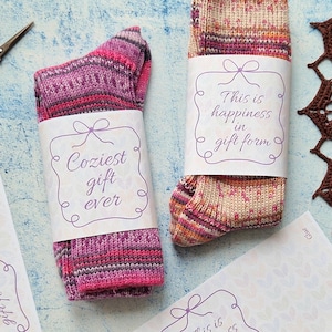 May include: Two pairs of knitted socks, one pink and the other beige, each with a label saying "Coziest gift ever" and "This is happiness in gift form." The socks are on a light blue surface with scissors and care instruction sheets.