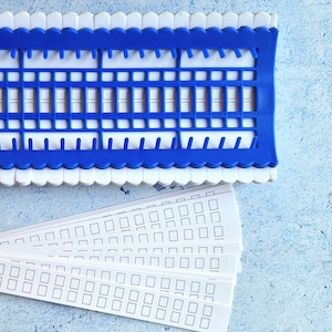 Può includere: Un telaio in plastica blu con pioli bianchi e un set di strisce in plastica bianca con quadrati stampati su di esse. Le strisce vengono utilizzate per la tessitura.