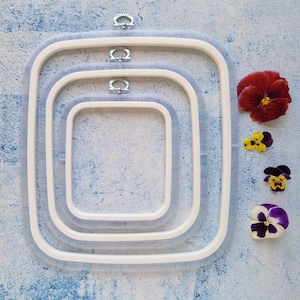 Puede incluir: Tres bastidores de bordado rectangulares encajados con marcos de plástico blanco y anillos interiores de color azul translúcido. Cada bastidor tiene un soporte de metal plateado para colgar en el centro superior. Cuatro flores de pensamiento coloridas están alineadas a la derecha.