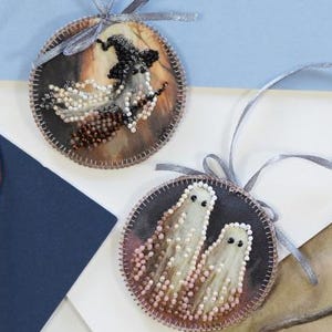 May include: Two round Halloween ornaments with beaded designs. One features a witch on a broom, the other two ghosts. Each ornament has a gray ribbon for hanging. The background includes a skull ribbon and a graveyard illustration.