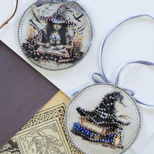 May include: Two round, beaded witch-themed ornaments with gray ribbons for hanging. One features a witch with a hat, the other a witch's hat on books. The ornaments have a light background with a brown paper and a drawing in the background.