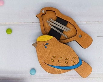 May include: A wooden bird-shaped needle minder with a blue and yellow design. The bird is open to reveal a compartment for storing needles.