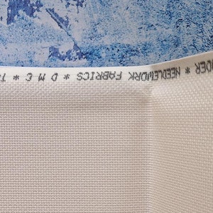 May include: Close-up of white cross-stitch fabric, with a visible woven texture. The fabric is rolled up, with the text "BRODER * NEEDLEWORK FABRICS * DMC * TOILES A BRODER * NEEDLEWORK" printed along the edge. The background is a textured blue.