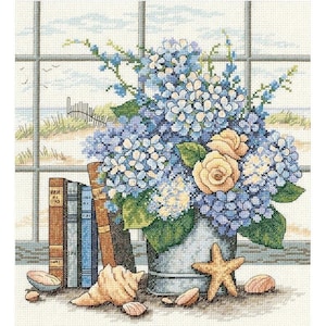 Op de afbeelding: Een stilleven met een boeket blauwe en witte hortensia's en gele rozen in een gegalvaniseerde metalen vaas. De vaas staat op een tafel met een stapel boeken en schelpen.