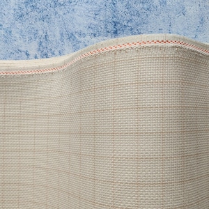 May include: Close-up of a beige woven fabric with a grid pattern. The fabric has a visible texture and a red and white stitched edge. The background is a mottled blue.