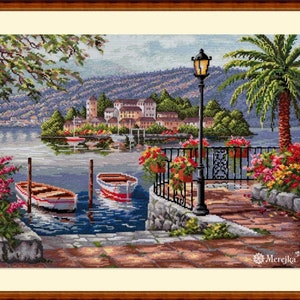 May include: A charming cross-stitch pattern depicting a picturesque lakeside scene with a quaint village nestled on a small island. The scene features a stone walkway lined with colourful flower boxes, a lamppost, and two boats docked at the shore. The water is a tranquil blue, and the sky is a soft, light blue.