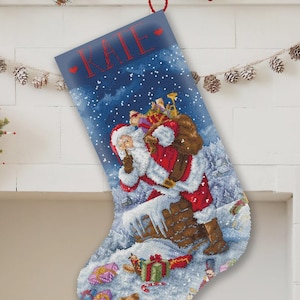 May include: A blue Christmas stocking featuring a cross-stitch design of Santa Claus holding a finger to his lips while standing on a snowy chimney. The name KATE is stitched in red at the top, flanked by two small red hearts against a starry sky.