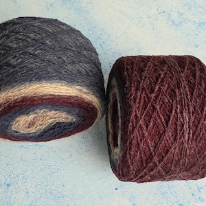 May include: Two cylindrical cakes of yarn. The left cake features a gradient of dark charcoal, cream, and deep burgundy. The right cake is a solid deep burgundy with a textured, fuzzy appearance. Both yarn cakes are wound into compact, round shapes.