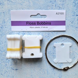 May include: A collection of white plastic floss bobbins, a bundle of floss, and a metal ring. The bobbins are labeled "Floss Bobbins" and measure 3.7 x 4 cm. The bundle of floss is secured with a yellow rubber band.