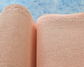 May include: Close-up of two sections of peach-colored linen fabric. The fabric has a woven texture and is folded over, with a line of stitching visible along the top edge. The background is a light blue color.