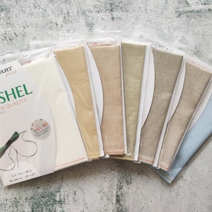 May include: A variety pack of Zweigart Cashel linen fabric in different colours, including white, beige, light brown, and blue. The fabric is 28 count and is made in Germany. The packaging includes the Zweigart logo and the text "Cashel Premium Quality".