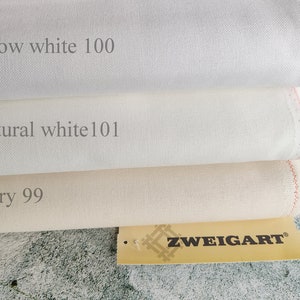 May include: Three different colours of Zweigart Aida fabric, from left to right: Snow white 100, Natural white 101, and Ivory 99. The fabric is folded and stacked on top of each other. A Zweigart label is visible in the bottom right corner.