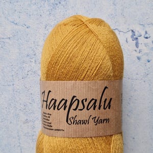 Puede incluir: Una madeja de hilo para chal Haapsalu de color amarillo dorado. El hilo está envuelto con una etiqueta de papel marrón. La etiqueta tiene el texto "Haapsalu Shawl Yarn" en una fuente manuscrita. El fondo es una superficie azul y blanca texturizada.