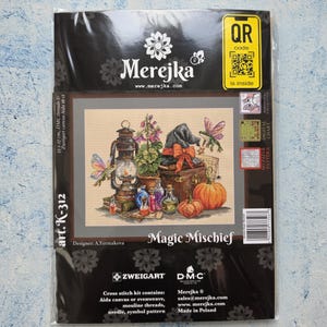 May include: A Merejka Magic Mischief cross stitch kit in plastic packaging. The cover features an illustration of a lantern, pumpkins, potion bottles, a witch hat, and a frog with wings. Text includes Merejka, Magic Mischief, and art. K-312.