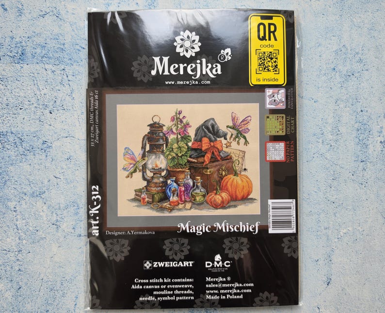 May include: A Merejka Magic Mischief cross stitch kit in plastic packaging. The cover features an illustration of a lantern, pumpkins, potion bottles, a witch hat, and a frog with wings. Text includes Merejka, Magic Mischief, and art. K-312.