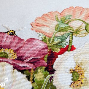 May include: Close-up of a cross-stitch embroidery featuring a bouquet of red, white, and pink poppies with green stems and leaves. A small bee is perched on one of the flowers.