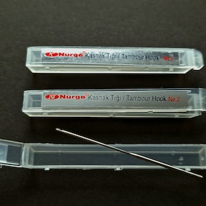 Könnte beinhalten: Zwei transparente Plastikboxen mit silbernen Tambourhaken darin. Die Boxen sind mit 'Nurgo Kasnak Tığı / Tambour Hook No. 1' und 'Nurgo Kasnak Tığı / Tambour Hook No. 2' beschriftet. Eine separate transparente Plastikbox enthält einen einzelnen silbernen Tambourhaken.