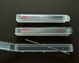 以下が含まれることがあります： 銀色のタムールフックが入った透明なプラスチックケースが2つ。ケースには「Nurgo Kasnak Tığı / Tambour Hook No. 1」と「Nurgo Kasnak Tığı / Tambour Hook No. 2」のラベルが貼られています。別の透明なプラスチックケースには、銀色のタムールフックが1つ入っています。