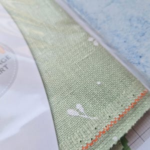 May include: Close-up of a light green fabric with white floral accents and an orange stitched edge. The fabric is partially visible, along with a white paper with text and a blue and white background.