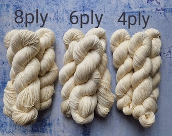 Könnte beinhalten: Drei Knäuel naturweißes Garn, beschriftet mit 8-fädig, 6-fädig und 4-fädig. Das Garn ist gedreht und in einem zopfartigen Muster angeordnet.