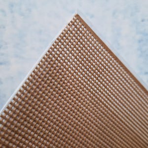 Könnte beinhalten: Nahaufnahme einer Ecke eines braunen, perforierten Materials, möglicherweise eine Tischset oder ein Bastelartikel. Das Material hat ein gitterartiges Muster aus kleinen, gleichmäßig verteilten Löchern. Der Hintergrund ist ein hellblaues und weißes, marmoriertes Muster.