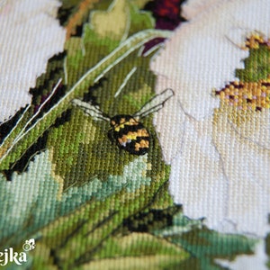 May include: Close-up of a cross-stitch embroidery depicting a white flower with a bee in the center. The bee is yellow and black with a brown body. The flower is surrounded by green leaves.